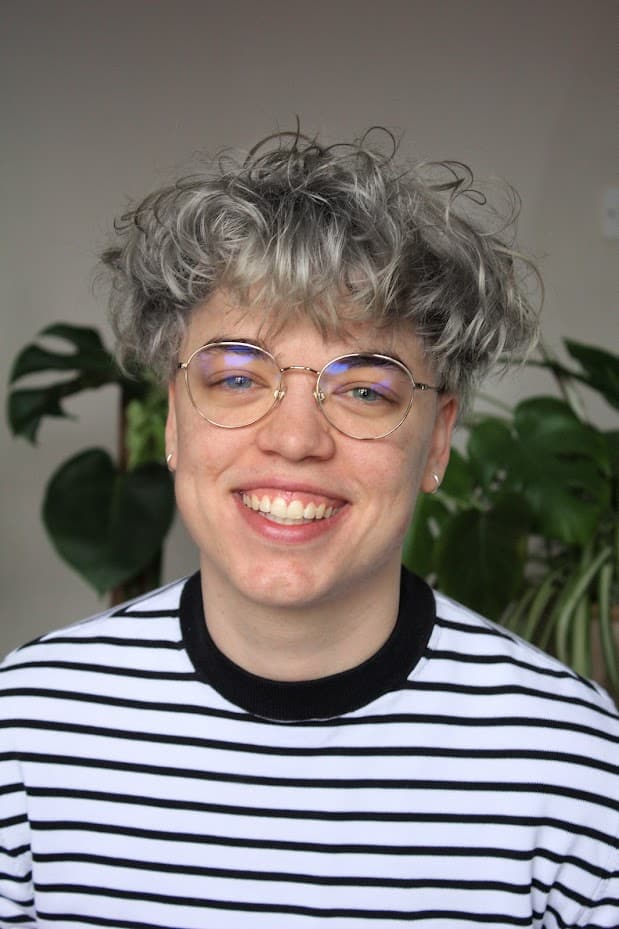 A person with curly gray hair, wearing glasses and a white and black striped shirt, smiling at the camera. A green plant is visible in the background.