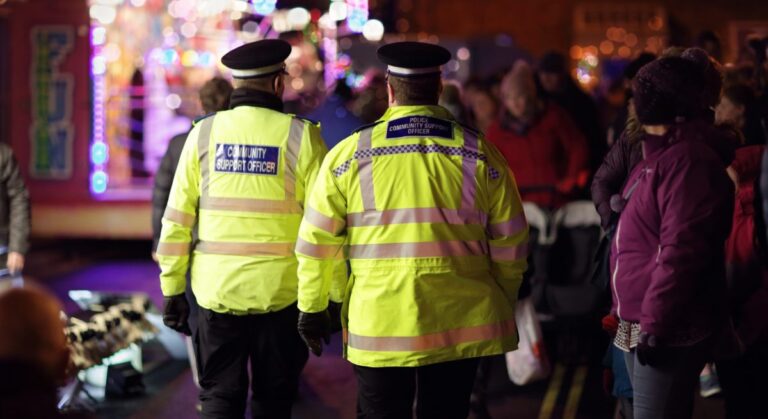 Two police officers in high-visibility jackets walk through a crowded, brightly lit street at night, surrounded by people and colorful lights.