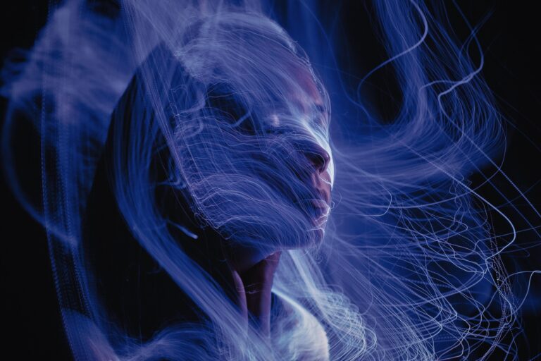 A person’s face is partially visible in low light, surrounded by swirling, glowing blue light trails against a dark background, creating a dreamy and surreal atmosphere.