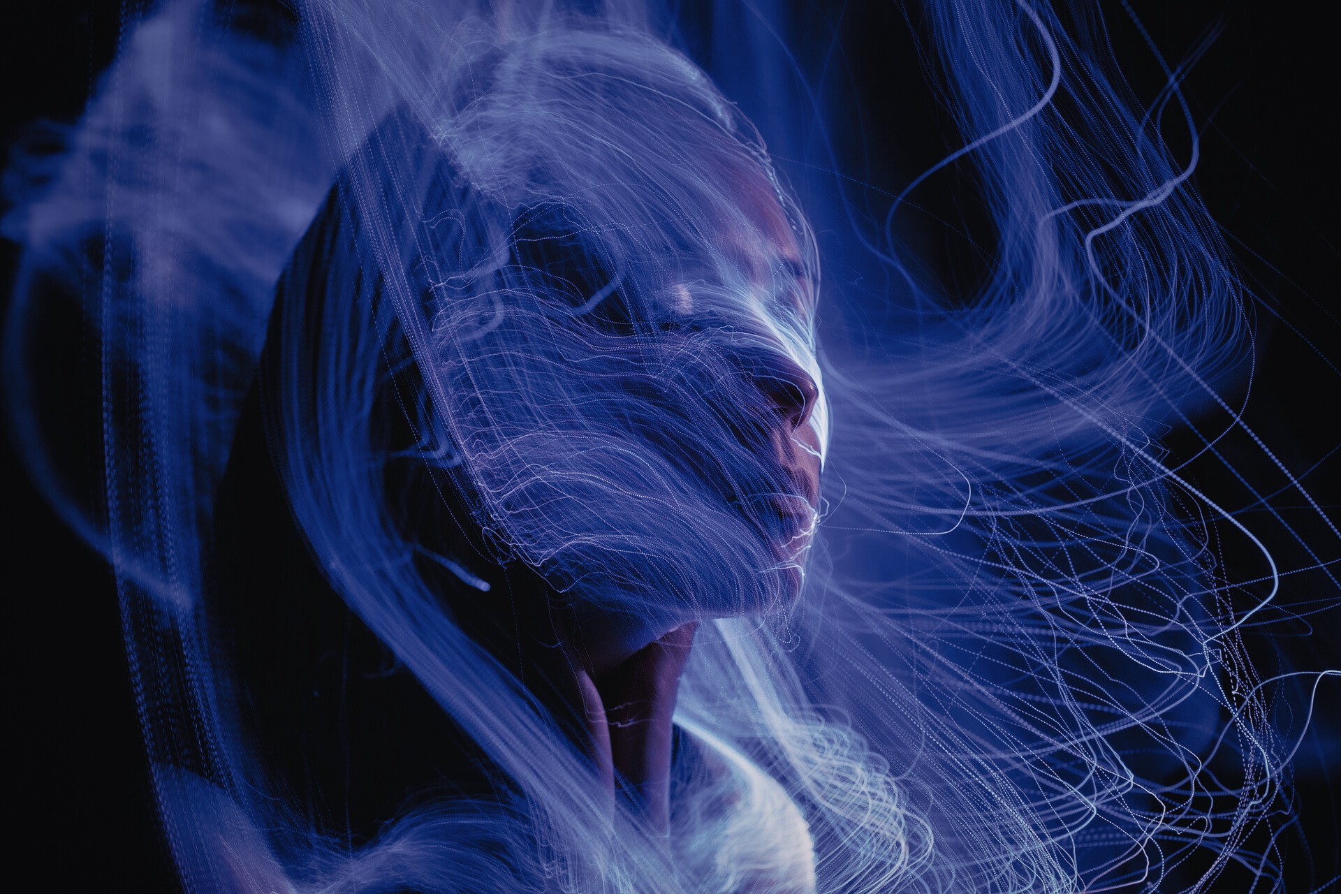 A person’s face is partially visible in low light, surrounded by swirling, glowing blue light trails against a dark background, creating a dreamy and surreal atmosphere.