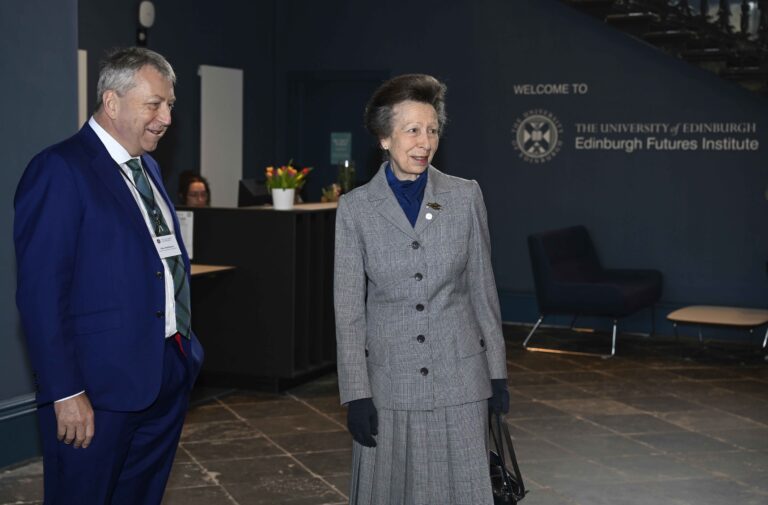 A woman in a gray suit and gloves stands next to a man in a blue suit inside the University of Edinburgh Futures Institute, with a reception desk and a welcoming sign in the background.