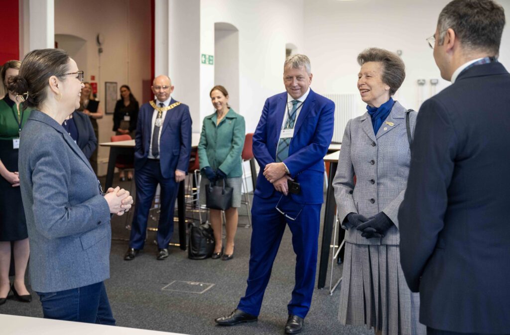 A group of formally dressed people stand indoors, smiling and conversing. A woman in a grey suit appears to be the center of attention, while others listen and interact in a well-lit, modern room.