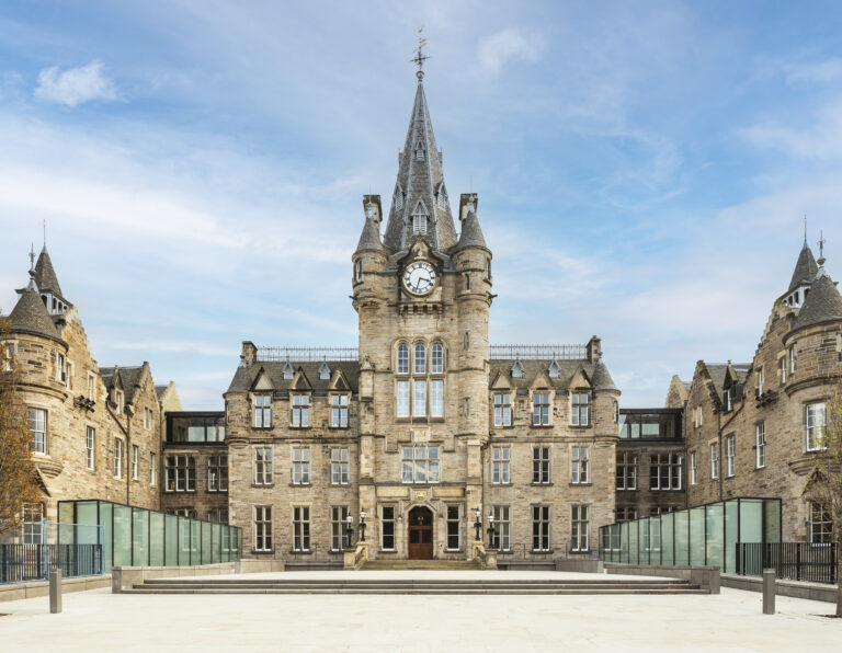 A large, historic stone building with a central clock tower and pointed turrets, surrounded by glass railings, under a blue sky. The area in front is a spacious, light-colored paved courtyard.