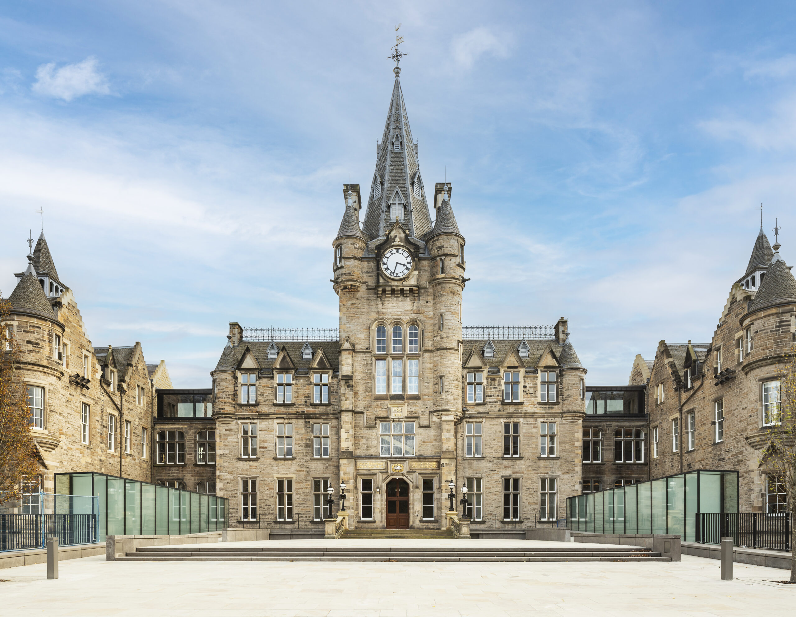 A large, historic stone building with a central clock tower and pointed turrets, surrounded by glass railings, under a blue sky. The area in front is a spacious, light-colored paved courtyard.