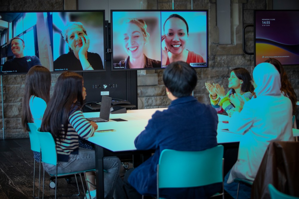 A group of people sit around a table in a modern meeting room, collaborating with three colleagues on video call shown on large screens. Some participants are smiling and clapping, suggesting a positive atmosphere.