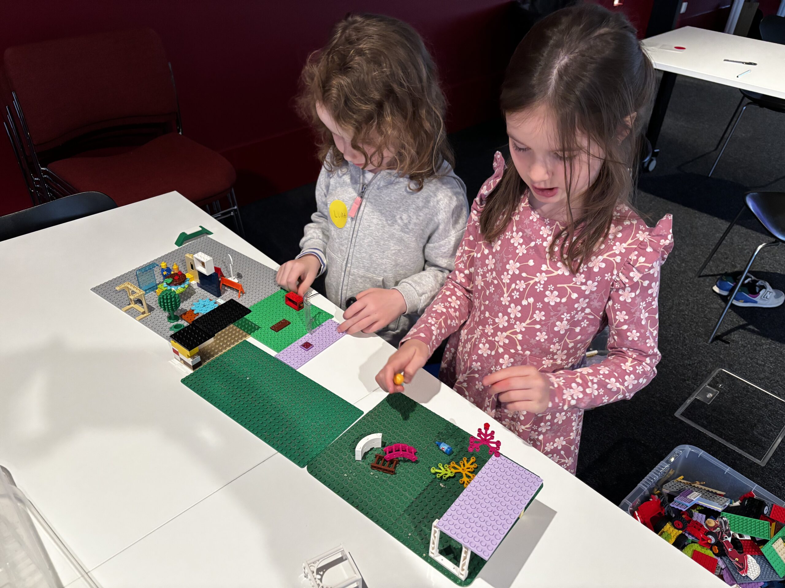 Two young children stand at a table, concentrating as they build and arrange colorful LEGO pieces on a green baseplate. A container of LEGO bricks is nearby, and the background includes chairs and tables.