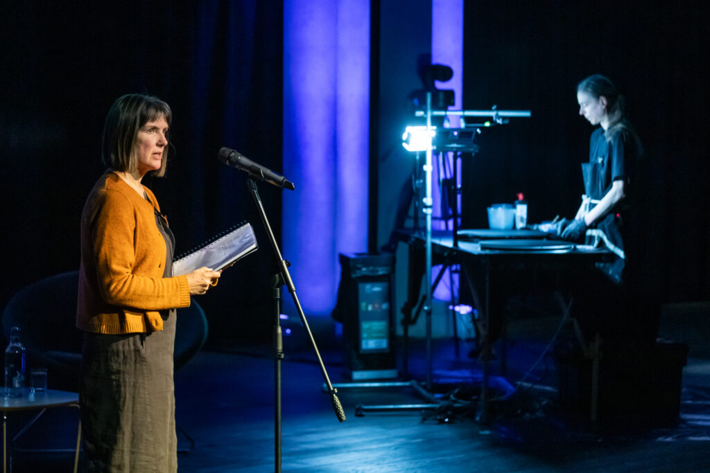A woman stands at a microphone holding papers, speaking on stage under blue lights. In the background, a technician works at a control desk with equipment and a bright lamp.