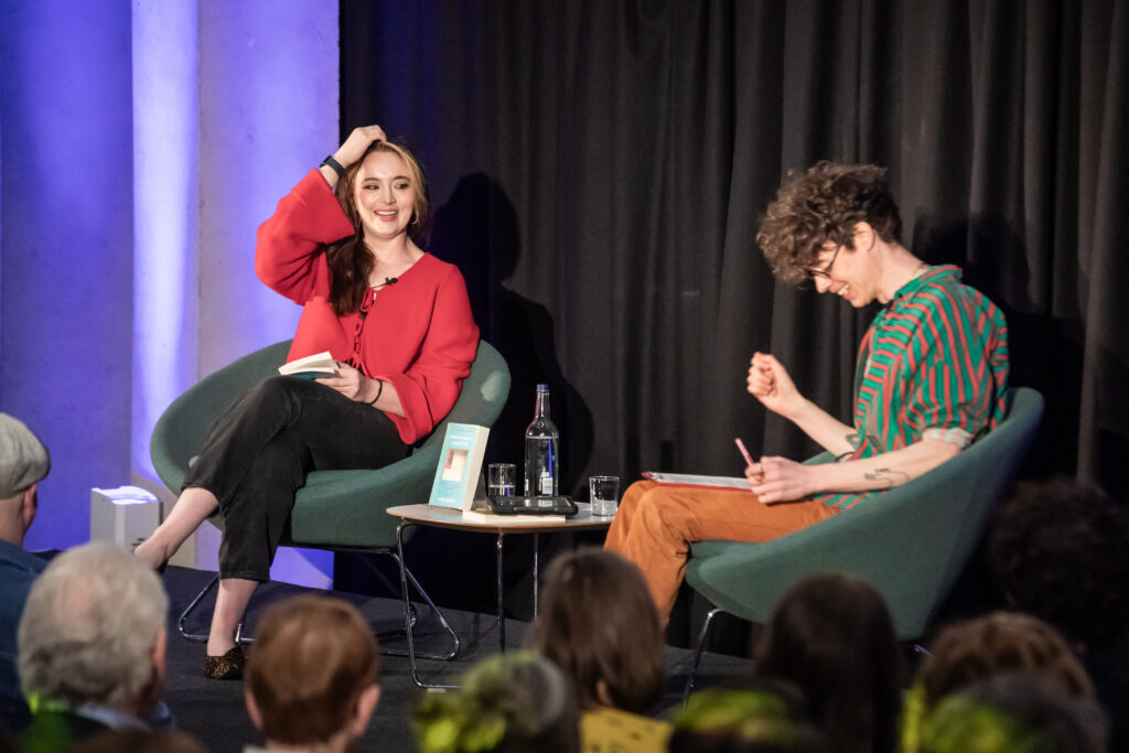 Two people sit on stage in modern chairs at a literary event. The woman on the left, smiling, holds a book and scratches her head. The man on the right takes notes. An audience watches them. A bottle and books are on the table.