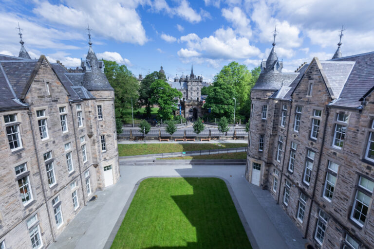 View through two historic stone buildings, framing a green lawn and a wide path, with a tree-lined road and a gothic-style gatehouse in the background under a blue sky with scattered clouds.