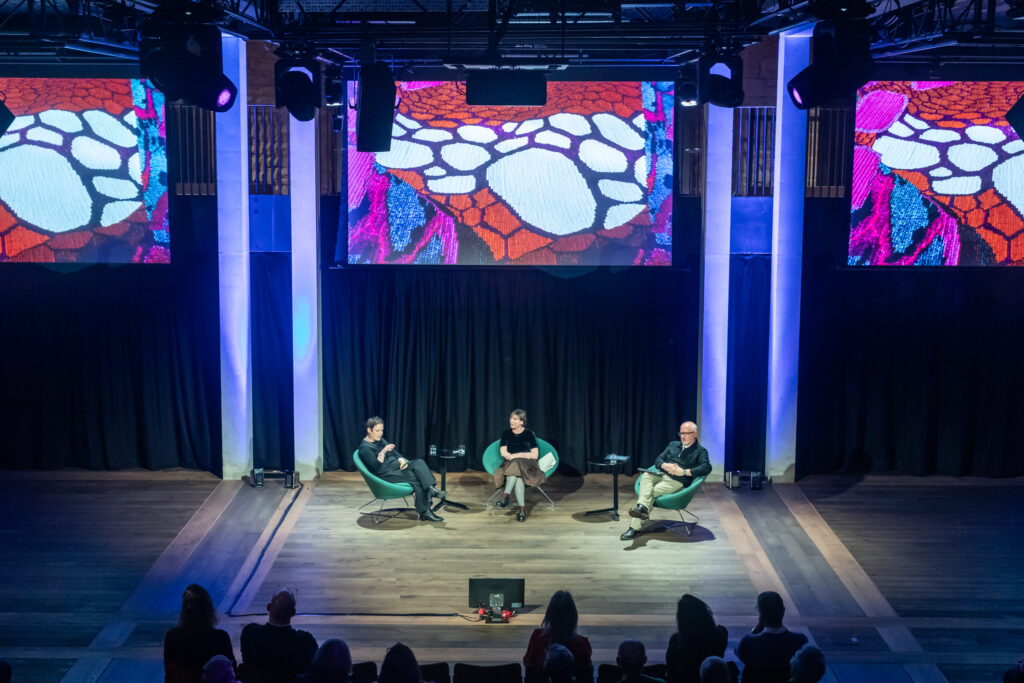 Three people sit on stage in chairs, engaged in a panel discussion. Colorful abstract visuals are displayed on two large screens above them. The audience watches from below in a dimly lit auditorium.