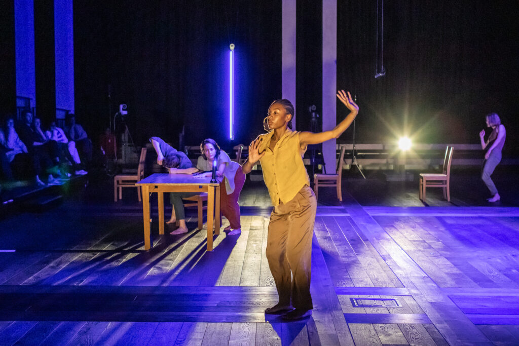 A performer in brown clothing dances under stage lights, with two people near a wooden table and others sitting or standing in the background. The scene is set on a dark stage with dramatic blue and yellow lighting.