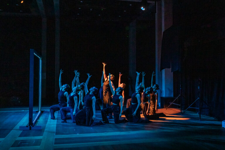 A group of dancers in dark costumes perform on a dimly lit stage, kneeling with arms raised toward blue and orange spotlights, creating dramatic shadows and a sense of unity.