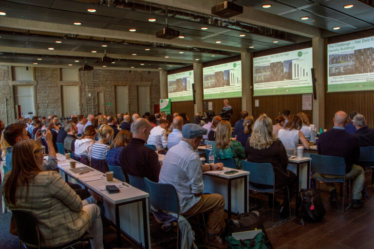A large group of people sit at tables in a conference room, facing a presenter and multiple screens displaying a presentation about net zero energy buildings. The room has stone walls and large windows.