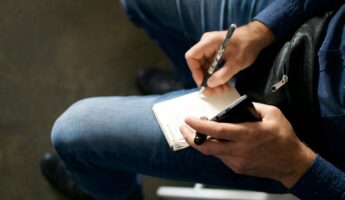 A person in blue jeans and a blue shirt sits and writes in a notebook with a pen, holding a smartphone in the other hand. Only their hands, lap, and part of the notebook are visible.