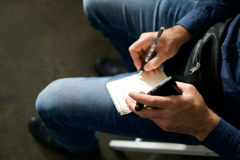 A person in blue jeans and a blue shirt sits and writes in a notebook with a pen, holding a smartphone in the other hand. Only their hands, lap, and part of the notebook are visible.