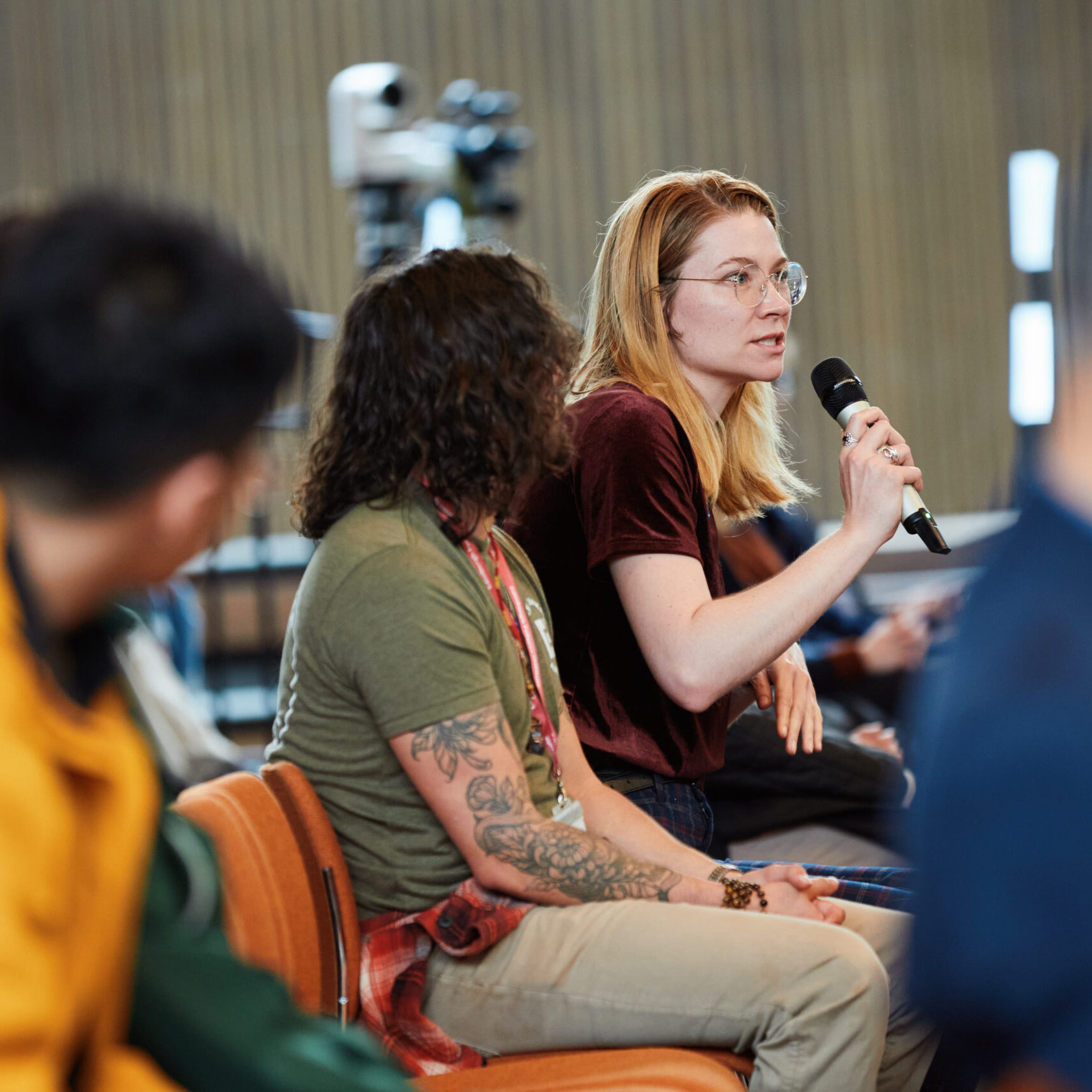 A woman with glasses speaks into a microphone while seated among a group of people at an indoor event or seminar. Others around her listen attentively.