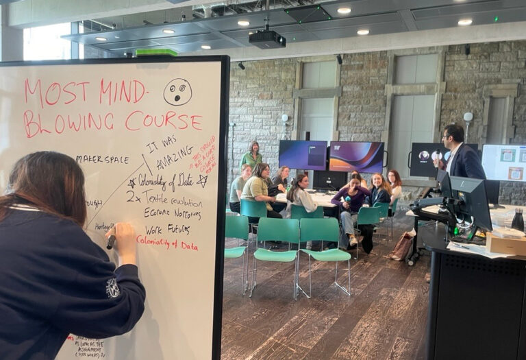 A person writes on a whiteboard labeled “Most Mind-Blowing Course” as a group of people sit in discussion around tables in a modern classroom with screens and a lecturer standing at the front.