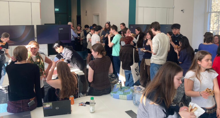A large group of people, mostly young adults, gather in a bright room with white walls, chatting, eating, and standing around tables with laptops and snacks. Sunlight streams through a window on the right.