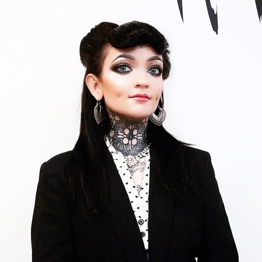 A woman with striking makeup, dark hair styled in vintage waves, a black suit, and a white shirt with black dots. She has heart-shaped earrings and detailed tattoos on her neck and chest, posing against a white background.