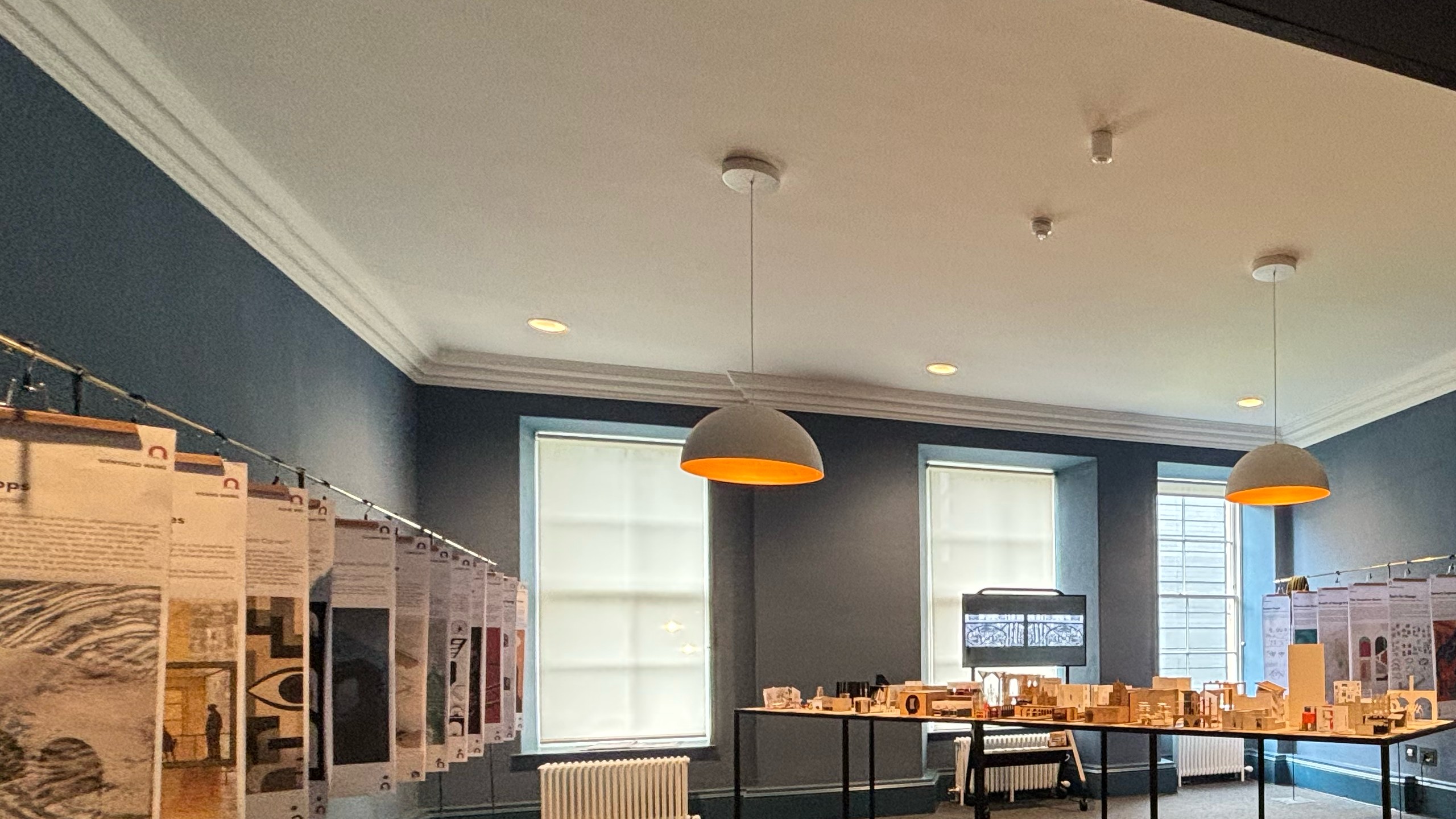 A room with blue walls displays hanging posters and architectural models on tables. Two round ceiling lamps and large windows provide natural light.
