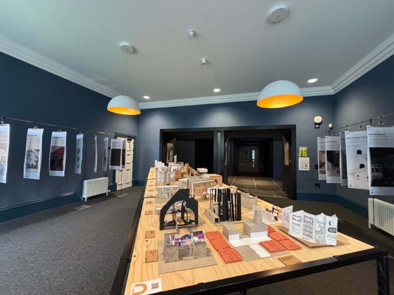 A gallery room with blue walls features a long table displaying architectural models and design books. Poster boards with text and images are hung along the walls. Two ceiling lights with orange shades illuminate the space.