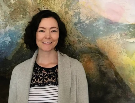Woman with curly dark hair, wearing a gray cardigan over a black and white striped top, stands smiling in front of a colorful abstract background.