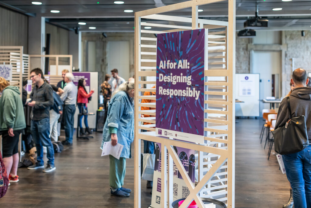 People attend an indoor event with informational booths. A prominent sign in the foreground reads “AI for All: Designing Responsibly.” The atmosphere is professional and collaborative.