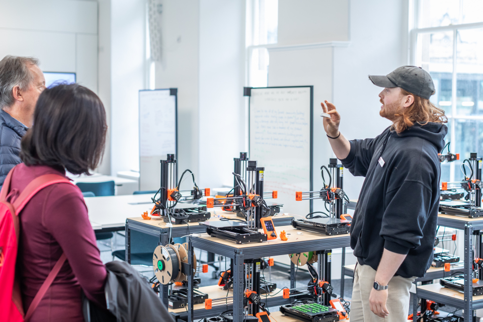 A man in a black cap gestures while explaining something to two people in a room filled with 3D printers and equipment. The setting appears to be a classroom or a workshop with large windows and whiteboards.