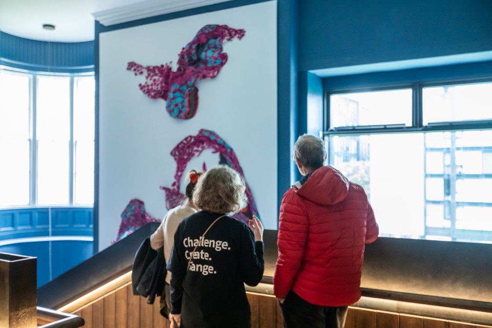 Two people stand indoors on a staircase, looking at a large abstract wall sculpture with pink and purple shapes. One person wears a red jacket, the other wears a black shirt that reads “Challenge. Create. Change.”.