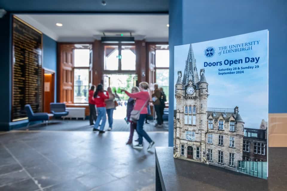 A poster for the University of Edinburgh’s Doors Open Day stands in focus indoors, while people in casual clothes enter the building through large open doors in the background.