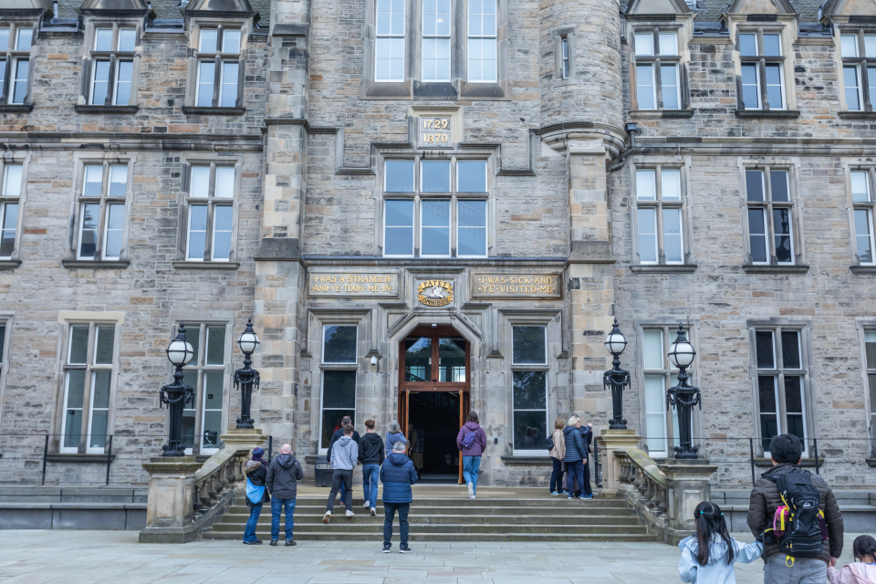 People stand and walk near the entrance of a large historic stone building with tall windows and ornate lamps. The building has signs above the door and a date plaque reading “1897” near the roof.