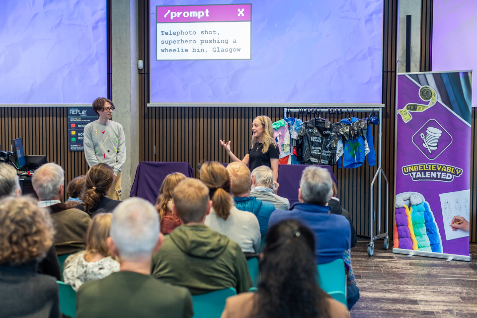 Two presenters stand at the front of a room, addressing an audience. A creative prompt is projected on a screen behind them. Colorful costumes hang on a rack, and a banner reads "UNBELIEVABLY TALENTED.