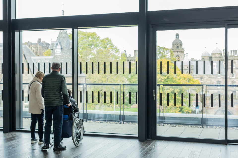 Three people, one in a wheelchair, stand by large glass windows overlooking a cityscape with historic buildings and trees on a cloudy day.