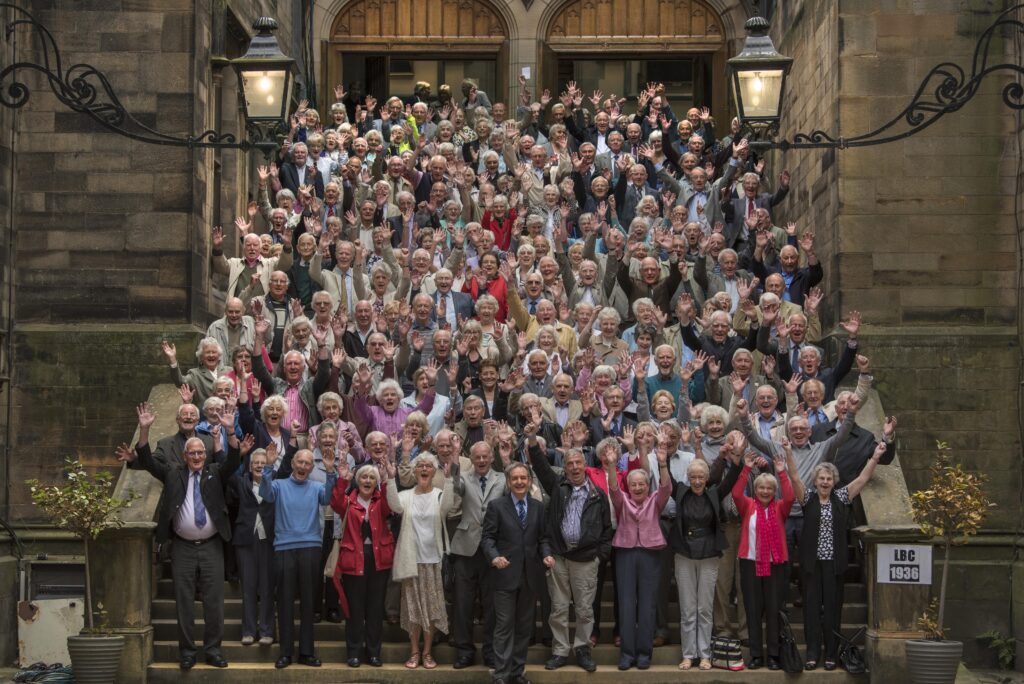A large group of elderly people pose together on the steps of a historic building, smiling and waving at the camera. The group appears cheerful, with many raising their hands, and the setting is outdoors in front of tall wooden doors.