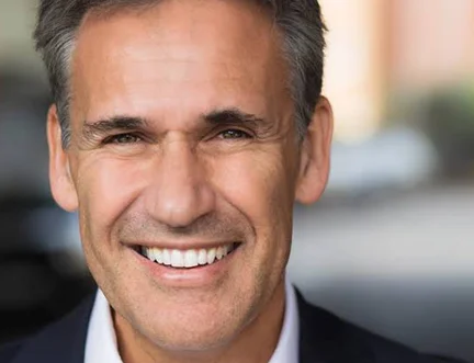 A middle-aged man with gray hair is smiling broadly, showing his teeth. He is wearing a dark suit jacket and is photographed outdoors with a blurred background.
