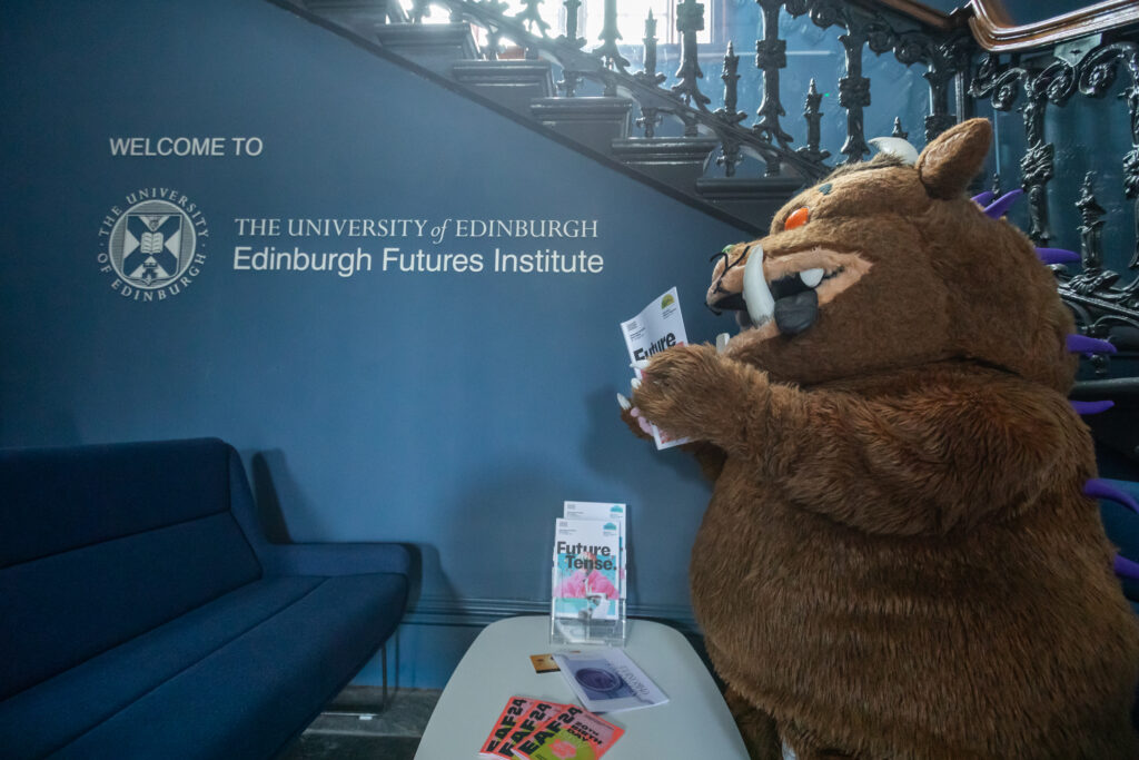 A person in a large brown monster costume stands in front of a wall sign reading "The University of Edinburgh, Edinburgh Futures Institute," holding a pamphlet near a table with brochures.