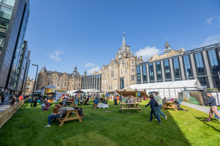 People gather on a grassy lawn with picnic tables, surrounded by historic and modern buildings under a sunny blue sky. Some are sitting, others walking, and a large tent is set up in the background.