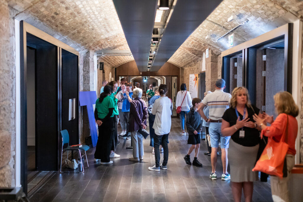 People gather and converse in a hallway with arched stone ceilings and wooden floors. Some stand in groups, while others walk or talk in pairs. The setting resembles a historic or museum-like building.