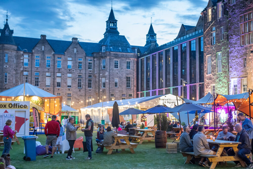 People sit and socialize at picnic tables in a lively outdoor festival area, surrounded by food stalls and festive lights, with historic stone buildings in the background at dusk.