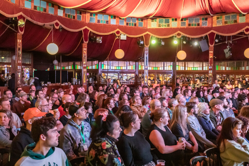 A large audience sits closely together under a colorful, ornate tent with red drapes and round hanging lights, attentively watching an unseen performance or presentation.