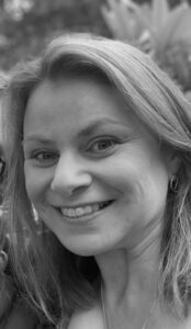A woman with light hair smiles at the camera in a close-up, outdoor setting. She is wearing hoop earrings and a necklace. The image is in black and white.