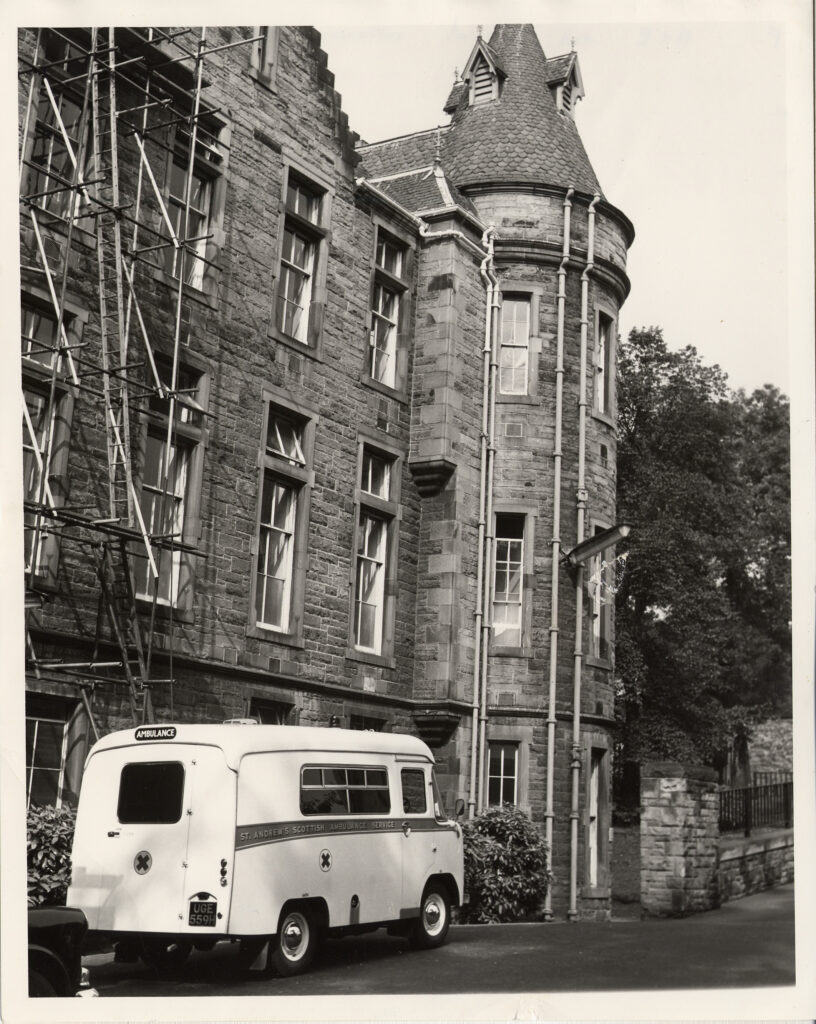 A black-and-white photo of a vintage ambulance parked outside a stone building with a turret and scaffolding on one side. Trees are visible in the background.