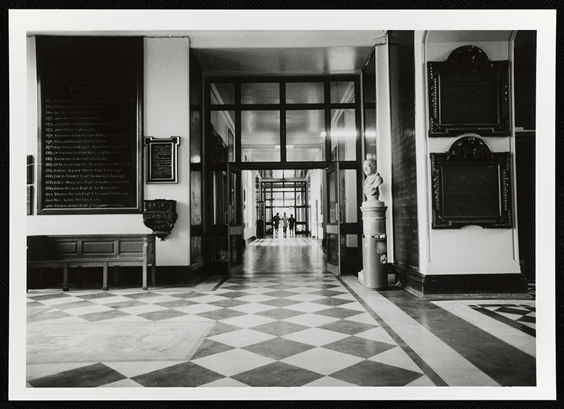 Black-and-white photo of a grand hallway with checkered floor tiles, plaques on the walls, a bust on a pedestal, and people visible in the distance through glass doors.