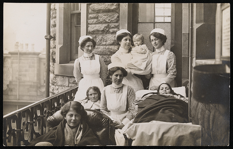 A vintage photo showing nurses and young female patients on a hospital balcony. Three nurses stand behind, one holding a baby, while four patients, two lying in beds, are in front of them, all dressed in early 20th-century attire.