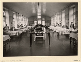 Black and white photo of a large hospital ward with rows of beds along both walls, patients lying in bed, nurses attending to them, and tables with flowers in the center aisle. Victorian-era setting.
