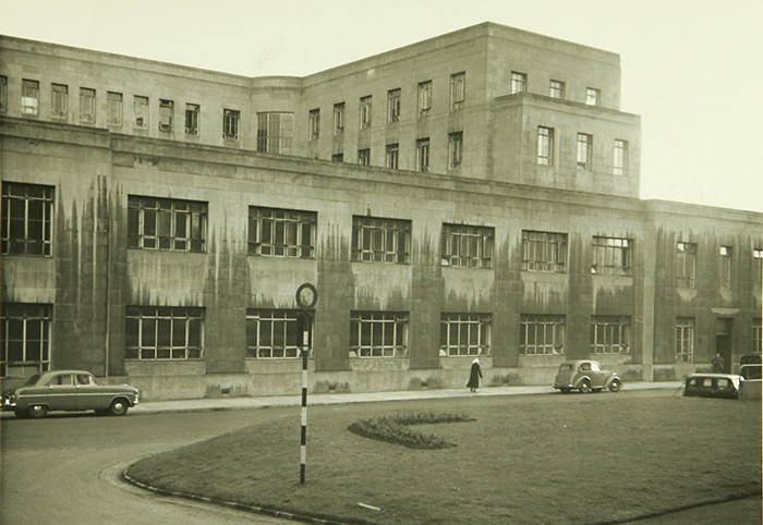 A black-and-white photo of a large, rectangular, three-story concrete building with many windows. Several vintage cars are parked along the curved road, and a person is walking on the sidewalk in front.