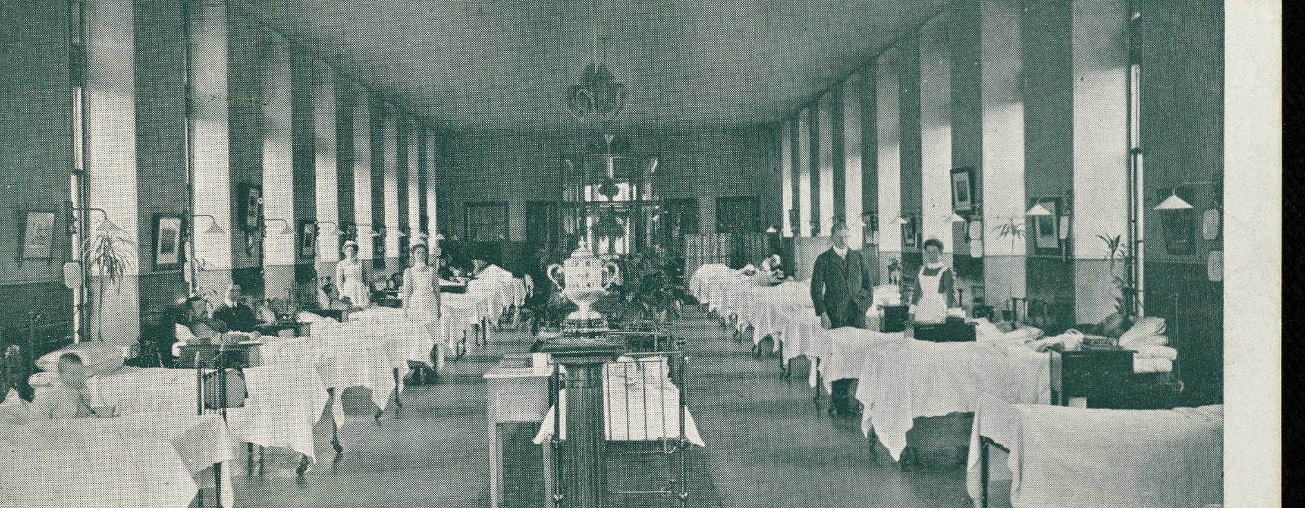 A vintage photograph of a long hospital ward lined with beds on both sides, nurses and patients present, tall windows along the walls, and a high ceiling with hanging lights.