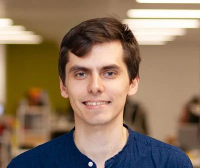 A young man with short dark hair, wearing a blue shirt, smiles at the camera in a brightly lit office with blurred people and desks in the background.