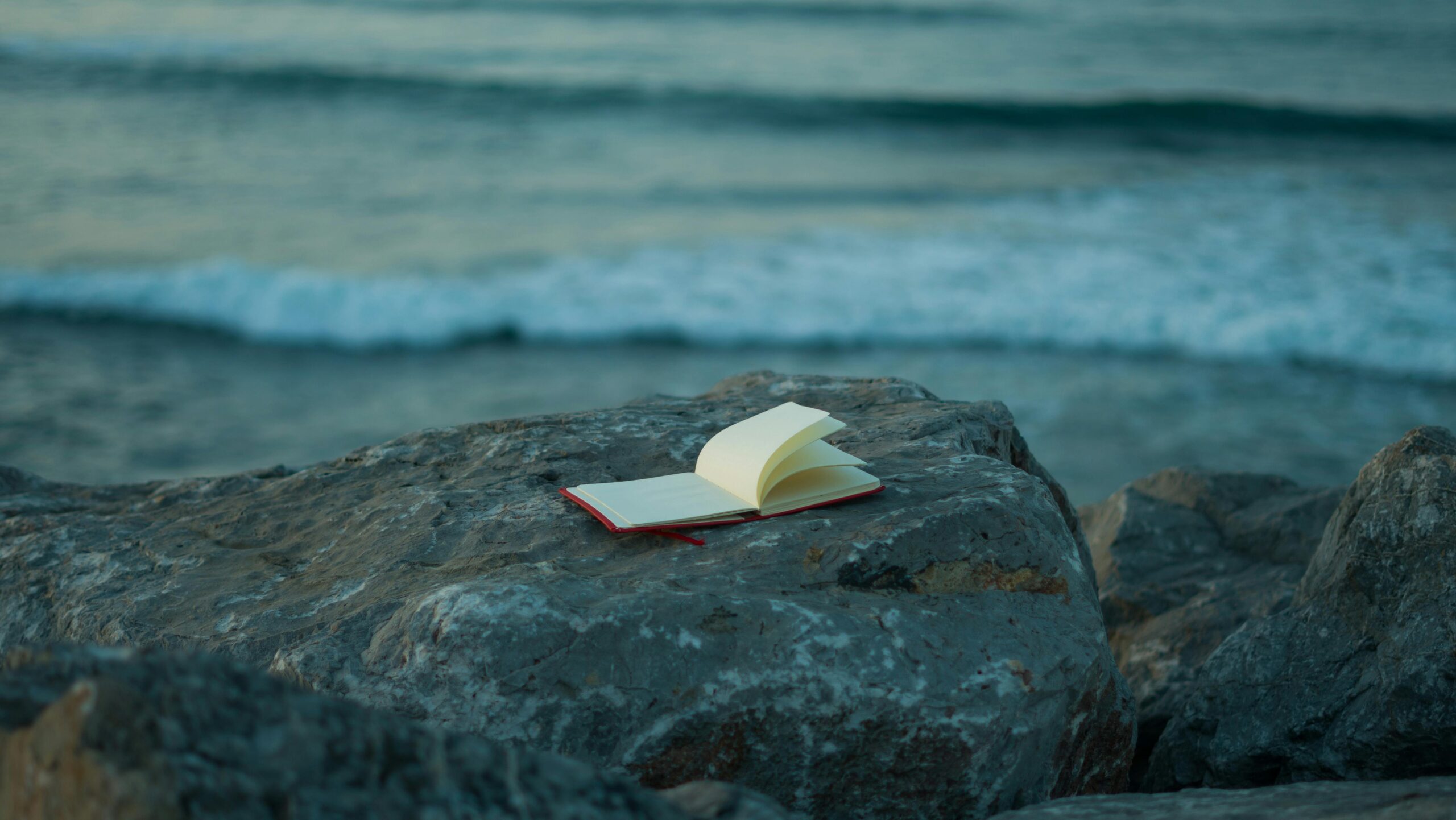 An open notebook with blank pages lies on a large rock by the seaside, with gentle ocean waves visible in the background.