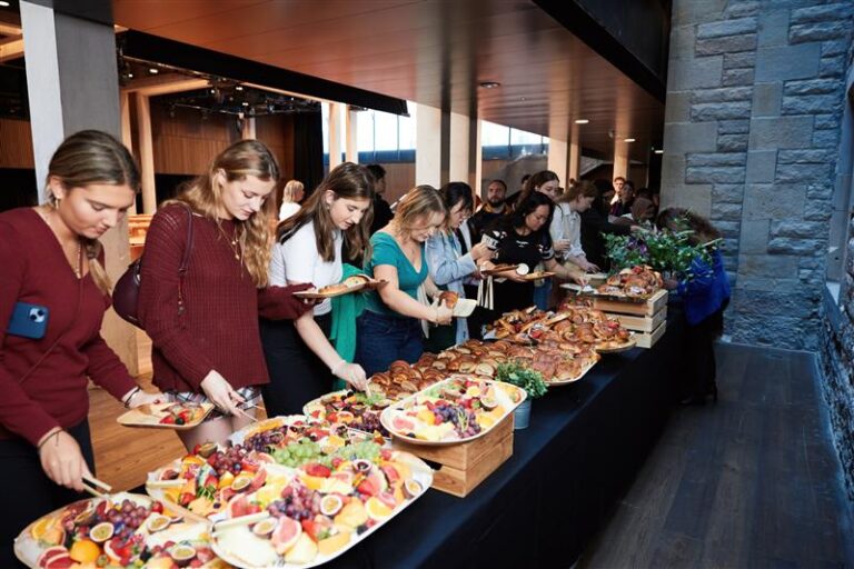 A group of people serve themselves fruit and pastries from a buffet table set up indoors, beside large windows and stone walls, during a social or corporate event.